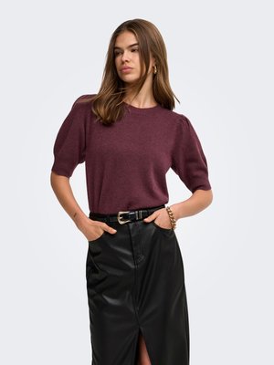 Femme portant un pull bordeaux à manches courtes et une jupe en cuir noire fendue, les mains dans les poches, et un bracelet en or au poignet gauche.