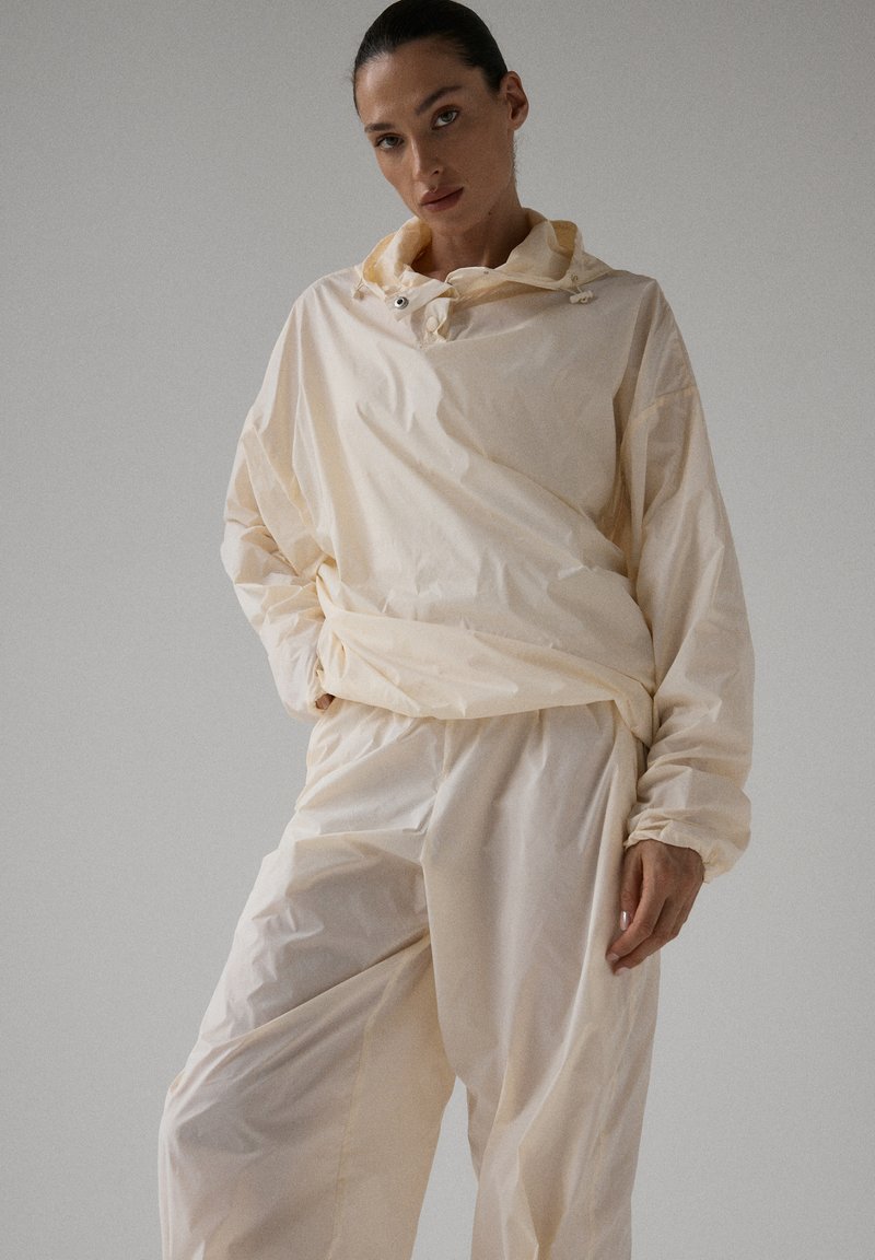 Kobieta w luźnej, jasnobeżowej kurtce przeciwwiatrowej z kapturem i dopasowanych spodniach, pozująca z jedną ręką w kieszeni na jednolitym tle.