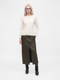 Femme blonde avec des lunettes portant un pull crème, une jupe en cuir vert foncé avec une fente à l'avant et des bottes à talons noires, debout devant un fond blanc.