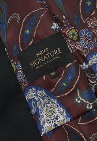 Burgundy-kuosiset kankaat, joissa on sinisiä ja vihreitä kukkakoristeita, mustalla etiketillä, jossa on kultakirjaimin teksti "NEXT SIGNATURE PREMIUM TAILORING" ja koko 40R.