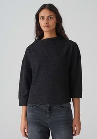 Schwarzes, strukturiertes Oberteil mit einer kastigen Silhouette, Dreiviertelärmeln und weitem Ausschnitt. Trägt dazu dunkelgraue Jeans. Subtile metallische Akzente.