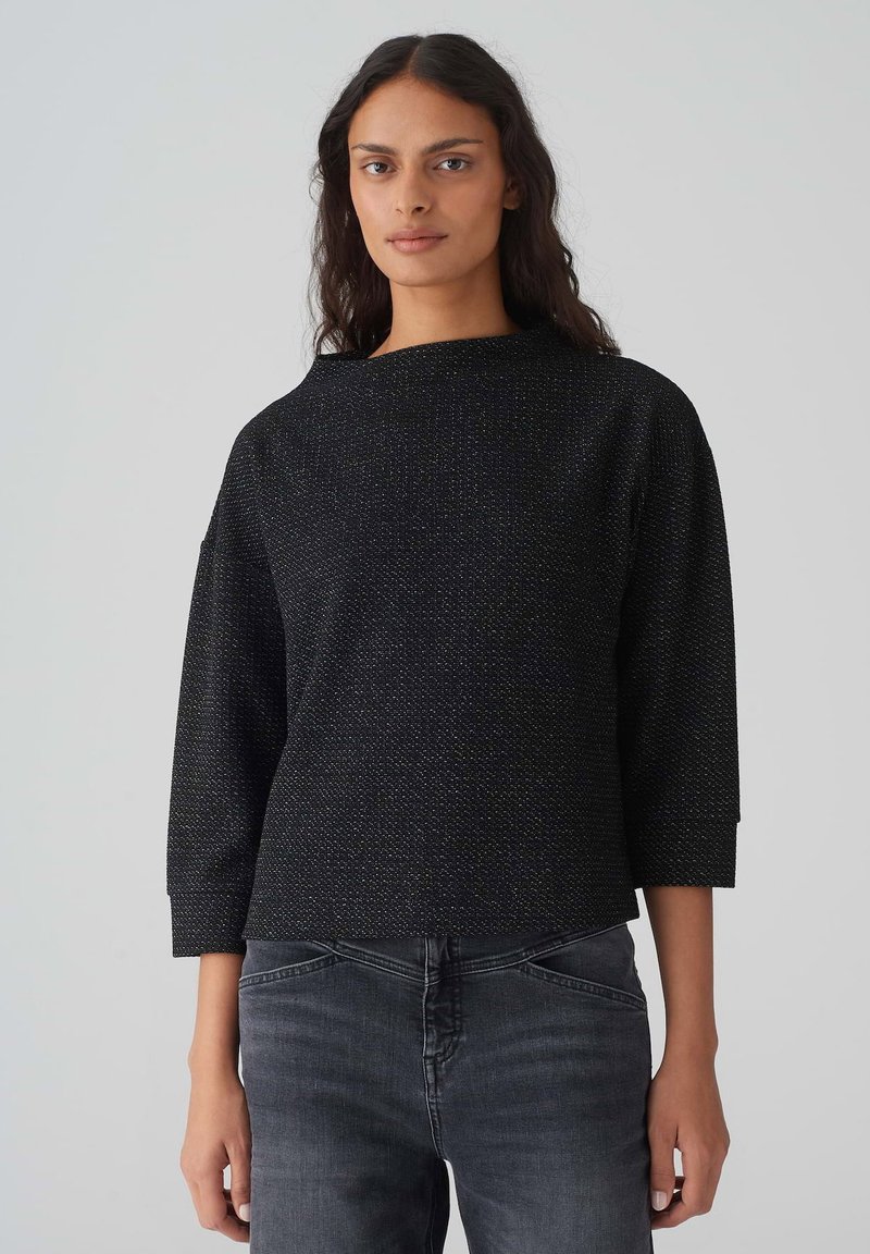 Schwarzes, strukturiertes Oberteil mit einer kastigen Silhouette, Dreiviertelärmeln und weitem Ausschnitt. Trägt dazu dunkelgraue Jeans. Subtile metallische Akzente.