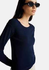 Granatowy sweter z długim rękawem, wykonany z miękkiego materiału dzianinowego, z okrągłym dekoltem i ściągaczem na dole, dopełniony czarnymi okrągłymi okularami przeciwsłonecznymi.