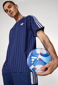 Jūrinio stiliaus, trumpomis rankovėmis, apvaliu iškirpimu ir logotipu sportinė marškinėliai su mėlynomis ir raudonomis dryžiais. Mėlynas ir raudonas raštuotas futbolo kamuolys laikomas dešinėje rankoje.