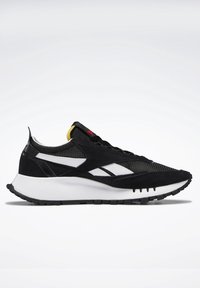 Baskets de sport noir et blanc avec des panneaux en mesh et en suède, une semelle blanche épaisse, et un petit détail jaune et rouge sur la languette.