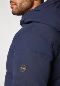 Mørkeblå pufferjakke med en teksturert utvendig overflate og et logo-patch på venstre arm. Har hette og quiltet design for isolasjon.