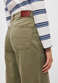 Παντελόνια σε χρώ�μα ελιάς με ευρύ μπατζάκι, πίσω τσέπες, υφασμένο ύφασμα με υφή, δερμάτινη ετικέτα επωνυμίας, συνδυασμένα με ριγέ πουκάμισο μακρυμάνικο.