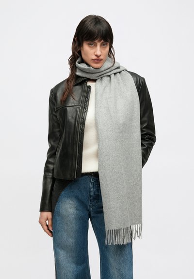 Écharpe en laine gris clair à bords frangés, drapée autour du cou d'une personne portant une veste en cuir noire et un pull crème, sur un fond uni.