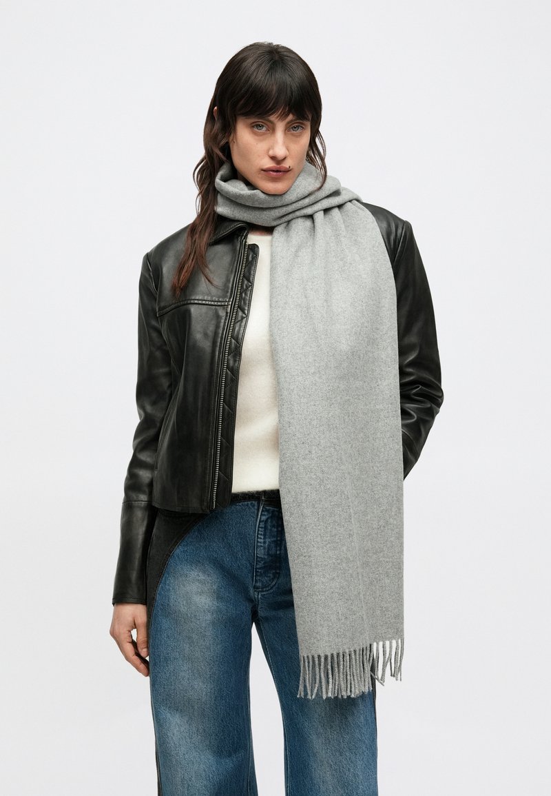 Sciarpa di lana grigio chiaro con bordi frangiati, drappeggiata intorno al collo di una persona con giacca di pelle nera e maglione crema, su sfondo semplice.