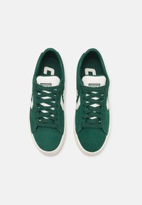 Green suede sneakers featuring a white rubber sole, rounded toe, and white accents on the sides and tongue. Laced design.