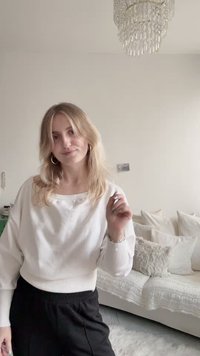 Λευκό φούτερ με ριμπ μανσέτες, σε συνδυασμό με μαύρο παντελόνι. Μακριά, κυματιστά ξανθά μαλλιά και ασημένια σκουλαρίκια κρίκοι. Απαλό, ουδέτερο φόντο.