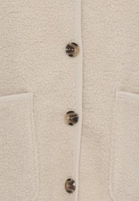 Section d'une veste en polaire texturée couleur crème avec trois boutons marbrés foncés et deux poches plaquées avant visibles.