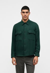 Zielona, teksturowana koszula z kołnierzykiem, dwiema kieszeniami na piersi i zapięciem na guziki z przodu. Długie rękawy z mankietami zapinanymi na guziki. Noszona na szarych spodniach.