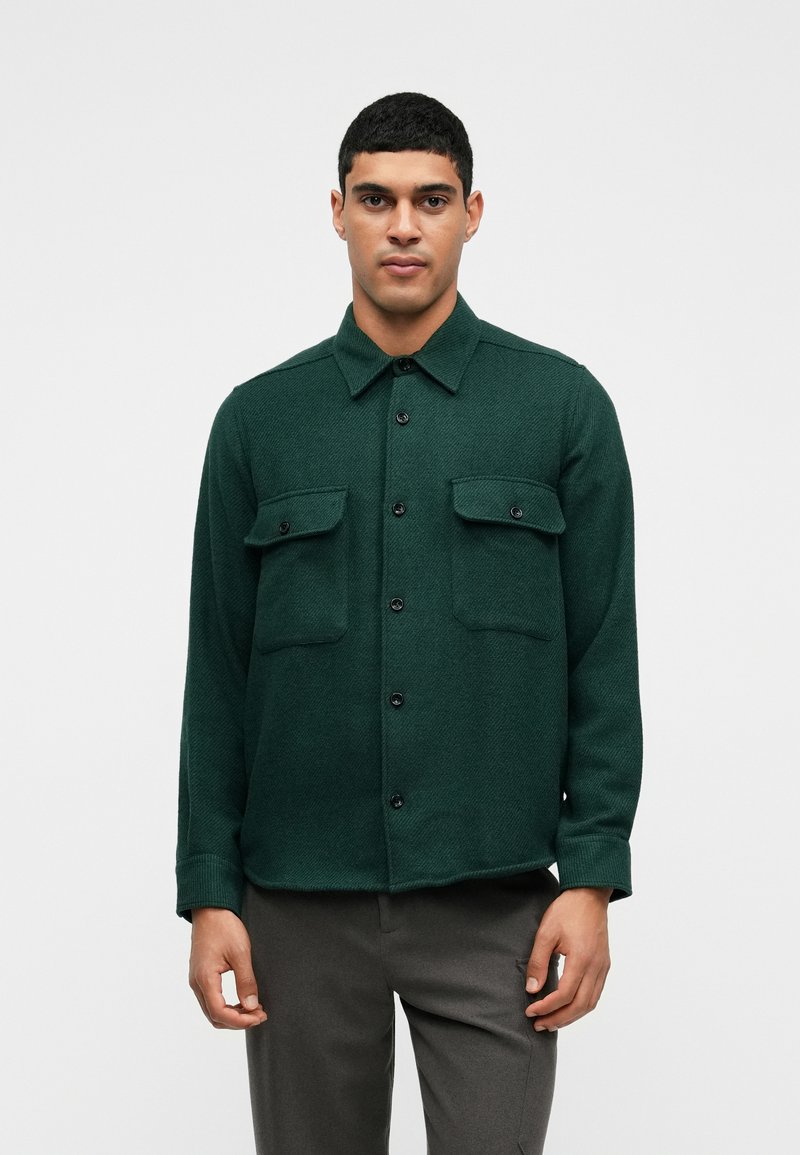 Zielona, teksturowana koszula z kołnierzykiem, dwiema kieszeniami na piersi i zapięciem na guziki z przodu. Długie rękawy z mankietami zapinanymi na guziki. Noszona na szarych spodniach.