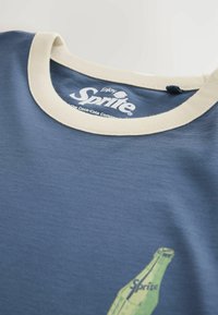 T-shirt bleu avec col crème arborant un petit motif de bouteille verte de Sprite et le texte imprimé en blanc "Enjoy Sprite" à l'intérieur de l'encolure.