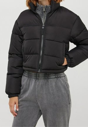 Personne portant une doudoune courte noire avec une fermeture éclair frontale et un pantalon gris décontracté à taille élastique, debout, une main dans la poche.