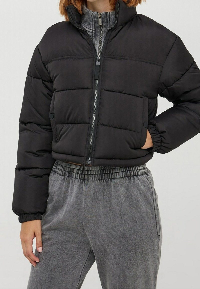Personne portant une doudoune courte noire avec une fermeture éclair frontale et un pantalon gris décontracté à taille élastique, debout, une main dans la poche.