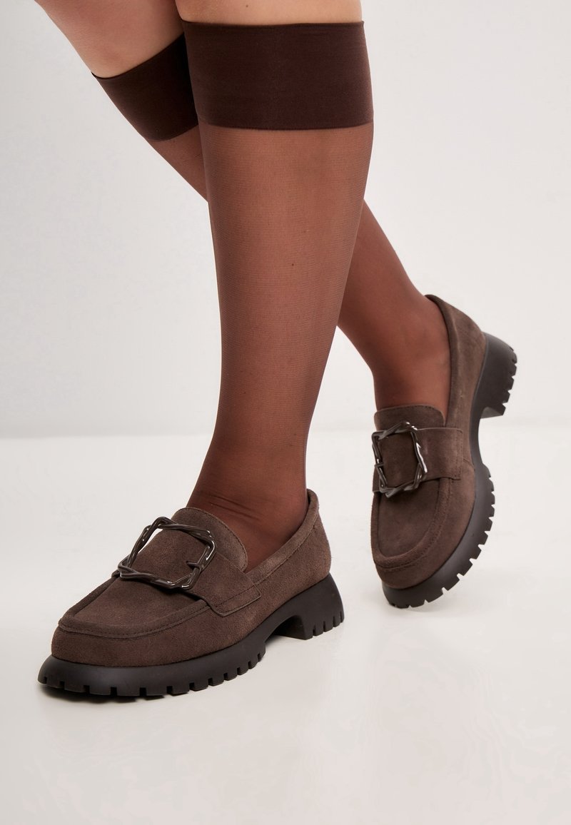 Mocassins en daim marron avec une semelle en caoutchouc noire épaisse et des détails en métal argenté. Portés avec des bas transparents marron jusqu'au haut de la cuisse.
