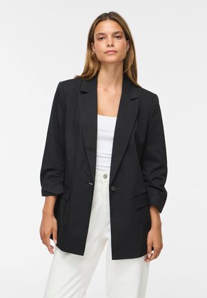 Kvinde iført en sort oversized blazer med optrukne ærmer, hvid top og hvide bukser mod en ensfarvet hvid baggrund.