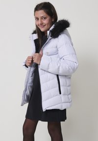 Chaqueta acolchada blanca con capucha de pelo sintético, con cierre frontal de cremallera, patrón de acolchado diagonal y un bolsillo en el pecho con un acento en forma de corazón dorado.