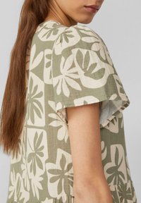 Frau mit langen braunen Haaren, die ein kurzärmliges grün-beiges Kleid mit floralem Muster trägt, ab der Schulter bis zur Mitte des Oberkörpers dargestellt.