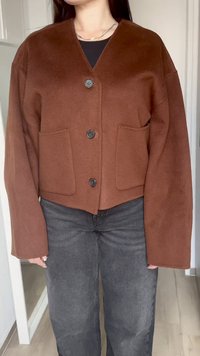 Brown cropped jacket made of soft fabric, featuring long sleeves, a v-neck, three buttons, and two front pockets. Worn with dark jeans.