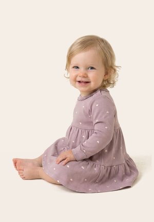 Mosolygó kisgyermek szőke hajjal mezítláb ül, hosszú ujjú levendulalila ruhát visel, kis fehér szívmintákkal, egyszínű háttér előtt.