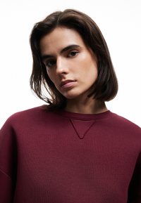 Sudadera burdeos con escote redondeado y costuras de hombro en relieve. Textura suave, con un sutil detalle en V en el cuello.