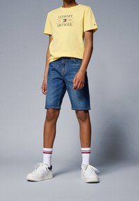 Żółta koszulka z krótkim rękawem z graficznym napisem "TOMMY HILFIGER", połączona z niebieskimi szortami dżinsowymi i białymi trampkami z niebieskimi akcentami oraz w pasiastymi skarpetkami.