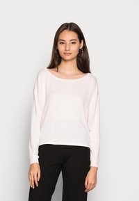 Camisa de manga longa rosa claro com um decote amplo e textura ribbed, combinada com calças pretas; modelo de pé contra um fundo simples.