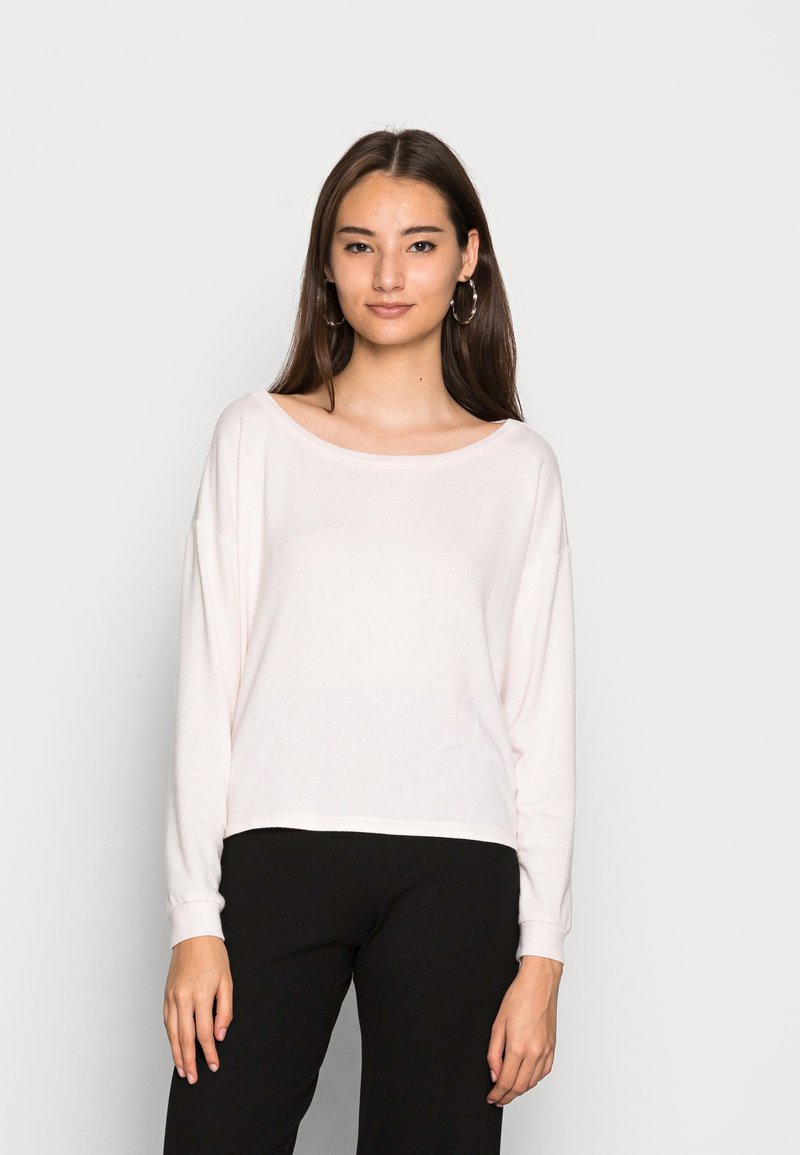 Camisa de manga longa rosa claro com um decote amplo e textura ribbed, combinada com calças pretas; modelo de pé contra um fundo simples.