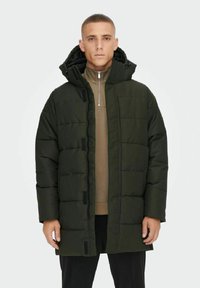 Veste rembourrée vert foncé avec capuche, présentant un design matelassé, fermeture éclair à l'avant et poches latérales. Portée par-dessus un haut marron.