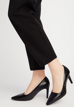 Zapatos de tacón alto de cuero negro con diseño de punta afilada, con una textura lisa y una silueta elegante. Usados con pantalones negros cortos.