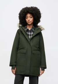 Groene parka-jas met een capuchon gevoerd met bont, grote voorkantenzakken en drukknoopsluitingen. Kenmerkt zich door een strak, mat oppervlak en een ontspannen pasvorm.