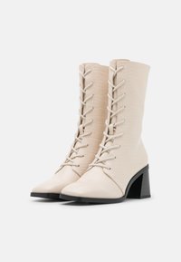 Beige Kunstleder Stiefeletten mit kroko-embossierter Textur, spitzer Zehenpartie, Schnürung vorne und einem massiven schwarzen Absatz.