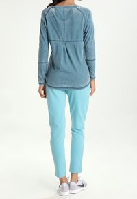Niebieska bluzka z długim rękawem, zaokrąglonym dekoltem i raglanowymi rękawami, zestawiona z jasnoniebieskimi spodniami i białymi trampkami. Gładka faktura materiału.