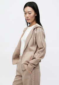 Φωτεινή μπεζ φούτερ με φερμουάρ μπροστά και κουκούλα, με υφασμένες πλαϊνές πινελιές. Συνδυάζεται με αντίστοιχο παντελόνι που έχει κέντημα με λογότυπο.