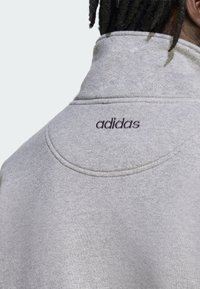 Felpa grigia chiaro con collo alto, caratterizzata da una finitura texturizzata sottile e dal logo "adidas" ricamato sul retro.