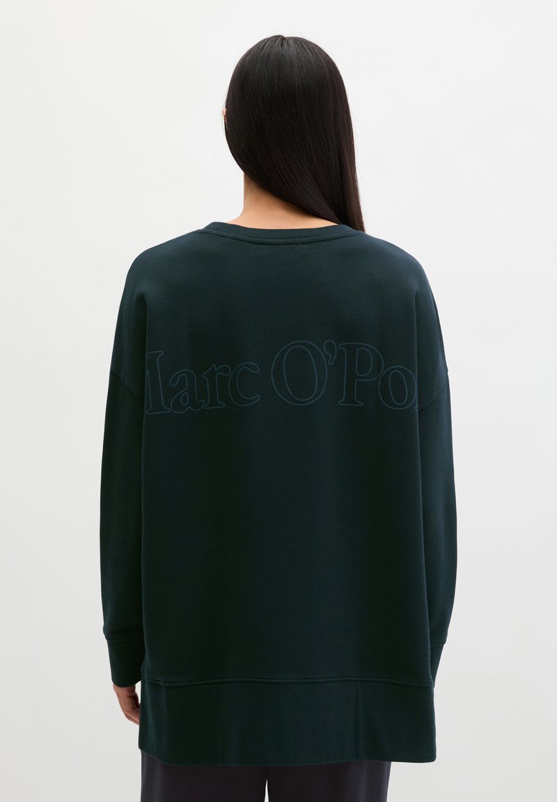 Femme aux longs cheveux foncés portant un sweat-shirt vert foncé oversize avec un discret texte "Marc O'Polo" au dos.