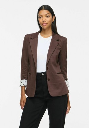 Femme portant un blazer marron avec doublure à pois aux poignets, un haut blanc et un pantalon noir, debout devant un fond blanc uni.