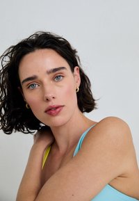 Retrato em close-up de uma pessoa com cabelo encaracolado e escuro, vestindo uma blusa azul e amarela, brincos de argola dourados e uma maquilhagem neutra.