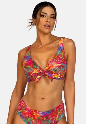 Kobieta ubrana w żywy, czerwony kwiatowy strój kąpielowy z wiązanym z przodu topem i dopasowanymi, wysokimi majtkami na jednolitym jasnym tle.