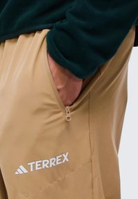 Beige Hosen mit einer Reißverschlusstasche und dem weißen "TERREX"-Logo. Der Stoff wirkt glatt mit einem subtilen Glanz und hat eine lockere Passform. Ein dunkelgrüner Ärmel ist teilweise sichtbar.