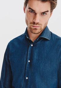 Ciemnoniebieska koszula dżinsowa o dopasowanym kroju, z długimi rękawami, kołnierzykiem zapinanym na guziki i białymi guzikami z przodu. Gładka tekstura.