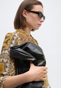 Γυναίκα με κοντά καστανά μαλλιά που φοράει μαύρα γυαλιά ηλίου και χρυσούς κρίκους στα αυτιά, κρατώντας μια διπλωμένη μαύρη δερμάτινη ζακέτα, φορώντας ένα κίτρινο-καφέ τοπ με σχέδια.