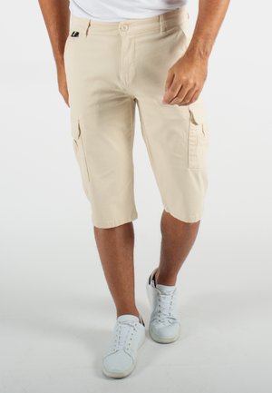 Shorts cargo beiges avec poches latérales, fabriqués en tissu léger. Présente une fermeture à bouton et une coupe décontractée. Associés à des baskets blanches.