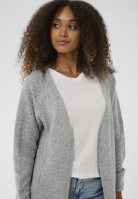 Világosszürke kötött kardigán, mintás textúrával az ujjakon. Fehér V-nyakú póló fölött viselve, kék farmerrel párosítva. Puha és kényelmes megjelenés.