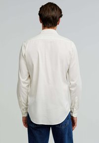 Weißes Langarmhemd mit Button-Down-Kragen, feiner Textur und abgerundetem Saum, kombiniert mit blauen Jeans, die eine lässige Passform zeigen.