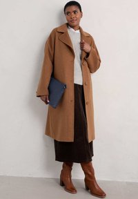 Barna gyapjú kabát, kihúzott gallérral, gombos záródással, bézs pulóverrel, barna kordbársony szoknyával és barna boka csizmával párosítva. Kék clutch táska.