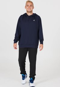 Marinblå hooded sweatshirt gjord av mjuk tyg, med en centrerad logotyp, kombinerad med svarta träningsbyxor och vita skor med blå accenter.